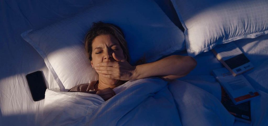 Woman shown in bed yawning with her devices and a book struggling to sleep