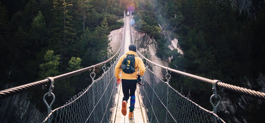 Hiker in a yellow jacket crosses a rope bridge across a ravine.