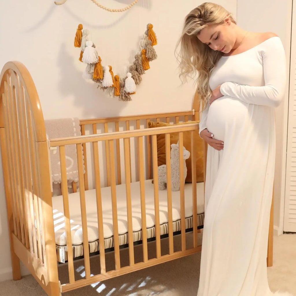 Pregnant woman cradling her baby bump wearing a white dress in front of her crib with a lala organic crib mattress in nursery